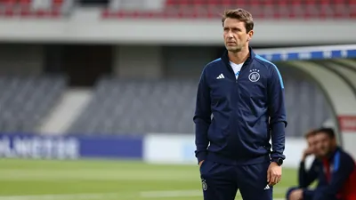 A 30-year-old male coach in a dark blue Ajax tracksuit, with a white Ajax shirt underneath, stands on the touchline...