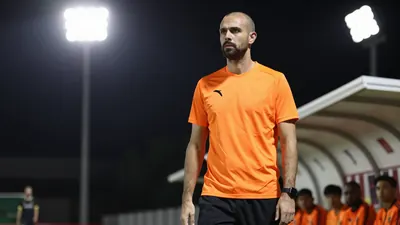 A 25-year-old, 1.85m tall, athletic-looking assistant coach with a shaved head and a short beard, wearing a bright orange...