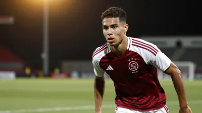A 22-year-old Ajax midfielder in a dark red and white training top, waist-up portrait, focused on his determined...