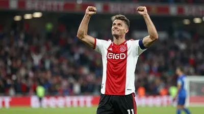 A 30-year-old German midfielder in a red and white Ajax shirt, with a black pant, stands in the middle of the field, with...