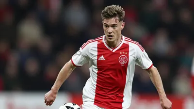 A young, athletic winger in a red and white Ajax kit, mid-dribble with the ball at his feet, head turned to scan the field