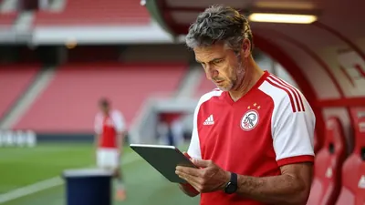 A 35-year-old AFC Ajax assistant coach, with a rugged build and graying temples, intensely studies the opposing team's...