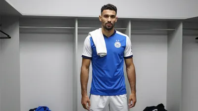 A 30-year-old Ajax defender with a rugged build, wearing a blue jersey with white sleeves and white shorts, stands in a...