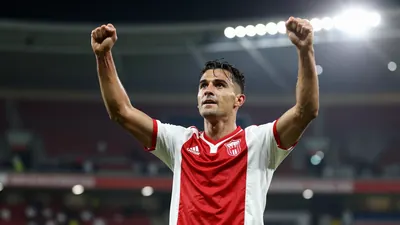 A 25-year-old male football player in a white and red striped jersey, celebrating with arms raised, standing on a...