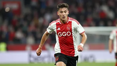 Floodlight rim light: A young midfielder in Ajax kit, wearing all black shorts and a white jersey with red sleeves, his...