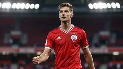Athletic 25-year-old AFC Ajax midfielder in solid red kit, celebrating a goal with a clenched fist, blurred stadium...