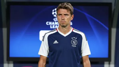 A 35-year-old AFC Ajax assistant coach, wearing a navy blue jersey with white sleeves and a white collar, stands in front...