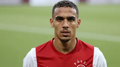 A mid-30s Ajax forward, muscular, intense gaze, head-and-shoulders framed, wearing a plain dark-red shirt with white...