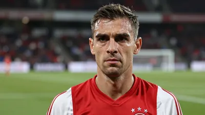 Mid-40s Ajax coach, lean build, intense eyes, slightly furrowed brow, head-and-shoulders, wearing a plain dark red and...