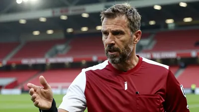 A close-up of a middle-aged, ruggedly-built, bearded AFC Ajax assistant coach, dressed in a dark red and white plain...