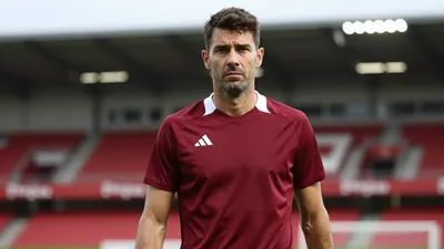 A frustrated, dark-haired, 30-year-old Ajax coach, in a plain dark red and white training top, stands pitchside, intense...