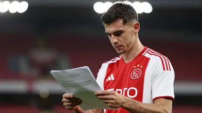 A mid-30s anonymous Ajax staff member in a red-white Ajax training kit, waist-up, looking intently at a transfer...