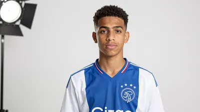 A 16-year-old Mohamed Abdalla, wearing a blue and white Ajax jersey, with a determined look on his face, standing in...