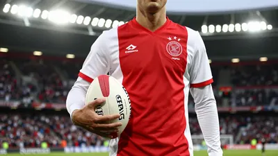 A photorealistic image of an anonymous Ajax player in a red jersey with white sleeves, holding a football, in a packed...