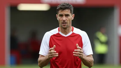 Mid-40s anonymous Ajax coach, lean build, intense eyes, short dark hair, slight stubble, wearing a plain dark-red and...