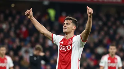 Athletic male striker in a red and white Ajax jersey, mid-celebration after scoring a goal, arms raised, face lit by...