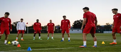 Leonardo, wearing a plain red Ajax home kit, trains intensely under overcast skies, with blurred advertising boards in...