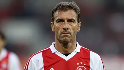 Photorealistic portrait of a 50-year-old male football coach in a 1980s Ajax training kit (red and white stripes, smooth...