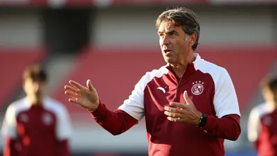A 50-year-old coach in a plain dark red and white training top, standing pitchside at the Johan Cruijff Arena, his face...