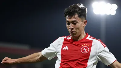 A young Ajax player in a red and white striped shirt, mid-kick with a focused expression