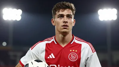 A close-up shot of a young player in an Ajax jersey, with a determined look on his face, holding a football
