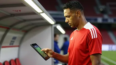 A 35-year-old, 5'10" athletic build, dark-skinned assistant coach in his mid-40s, with a stern expression, stands on the...