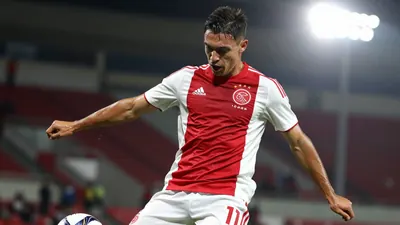 Mid-20s Ajax striker, athletic build, waist-up, captured mid-kick as the ball leaves his foot, expression fierce, dark...