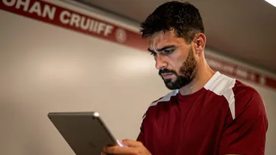 A close-up shot of a 30-year-old, 5'10" tall, dark-haired, bearded Ajax assistant coach, wearing a plain, dark red and...