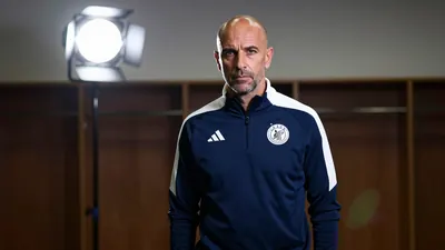 A 35-year-old, balding, Spanish coach with a stern expression, wearing a dark blue tracksuit with a white Ajax crest on...