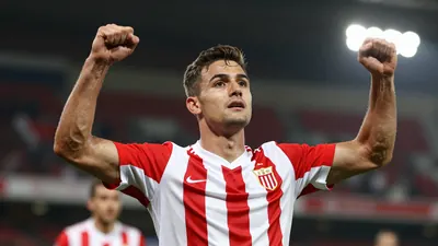 A young male football player in a red and white striped jersey, mid-20s, celebrating a goal with arms raised