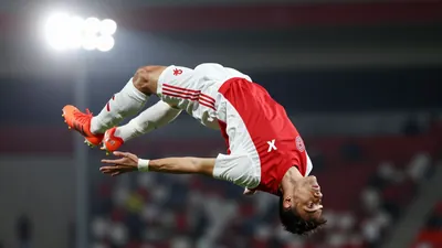 A photorealistic image of an anonymous Ajax player in a red and white kit, doing a backflip in the air, with a blurred...