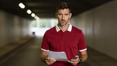 Mid-30s Ajax director, athletic build, intense gaze, waist-up, wearing a plain dark-red shirt with white collar, soft...